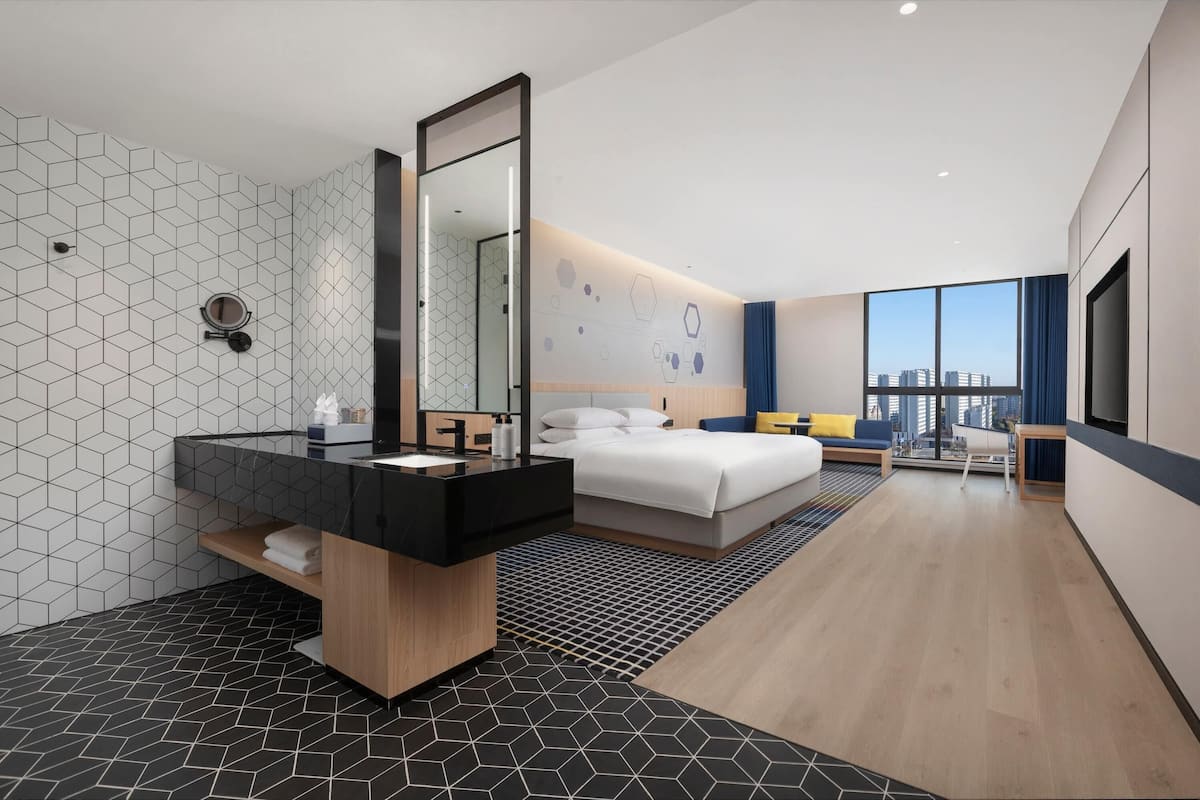Ein modernes Hotelzimmer mit einem großen Bett, einem Badezimmer mit schwarzem Waschtisch, einem großen Spiegel und Blick auf die Stadt durch das Fenster.