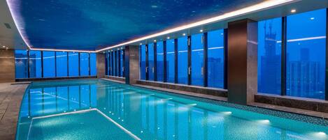 A long indoor swimming pool with clear blue water, surrounded by large windows offering city views.