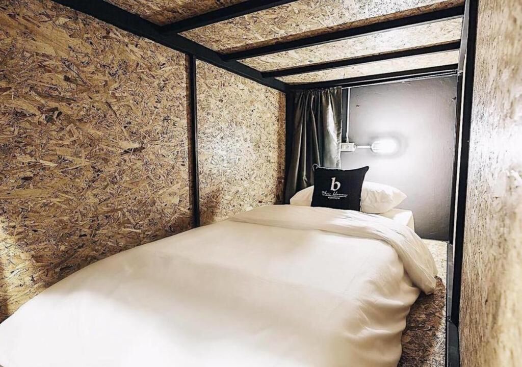 A single bed with a white mattress and pillows, a black pillow with a white logo, a wooden textured wall, and a door with a light fixture.