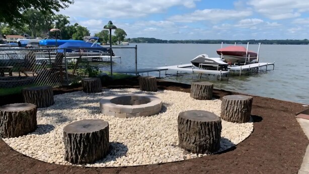 A lakeside deck with a circular seating area surrounded by wooden stumps and white pebbles, overlooking a calm lake with boats docked nearby. - Pistakee Lake-Front Retreat (Fox Lake)