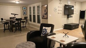 A living room with a black leather chair, a white table, a television, and a dining area with a bar stool. - Pistakee Lake-Front Retreat (Fox Lake)