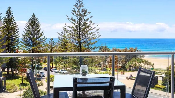 Un balcon avec une table et des chaises donnant sur une plage bordée d’arbres, sous un ciel dégagé.