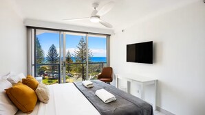Une chambre d’hôtel moderne dotée d’un grand lit, d’un bureau, d’une chaise, d’une télévision à écran plat et d’un balcon offrant une vue sur l’océan et la végétation.
