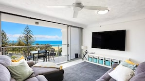 Un salon moderne doté d’un grand téléviseur, de canapés gris, d’une table basse et d’un balcon offrant une vue sur l’océan.