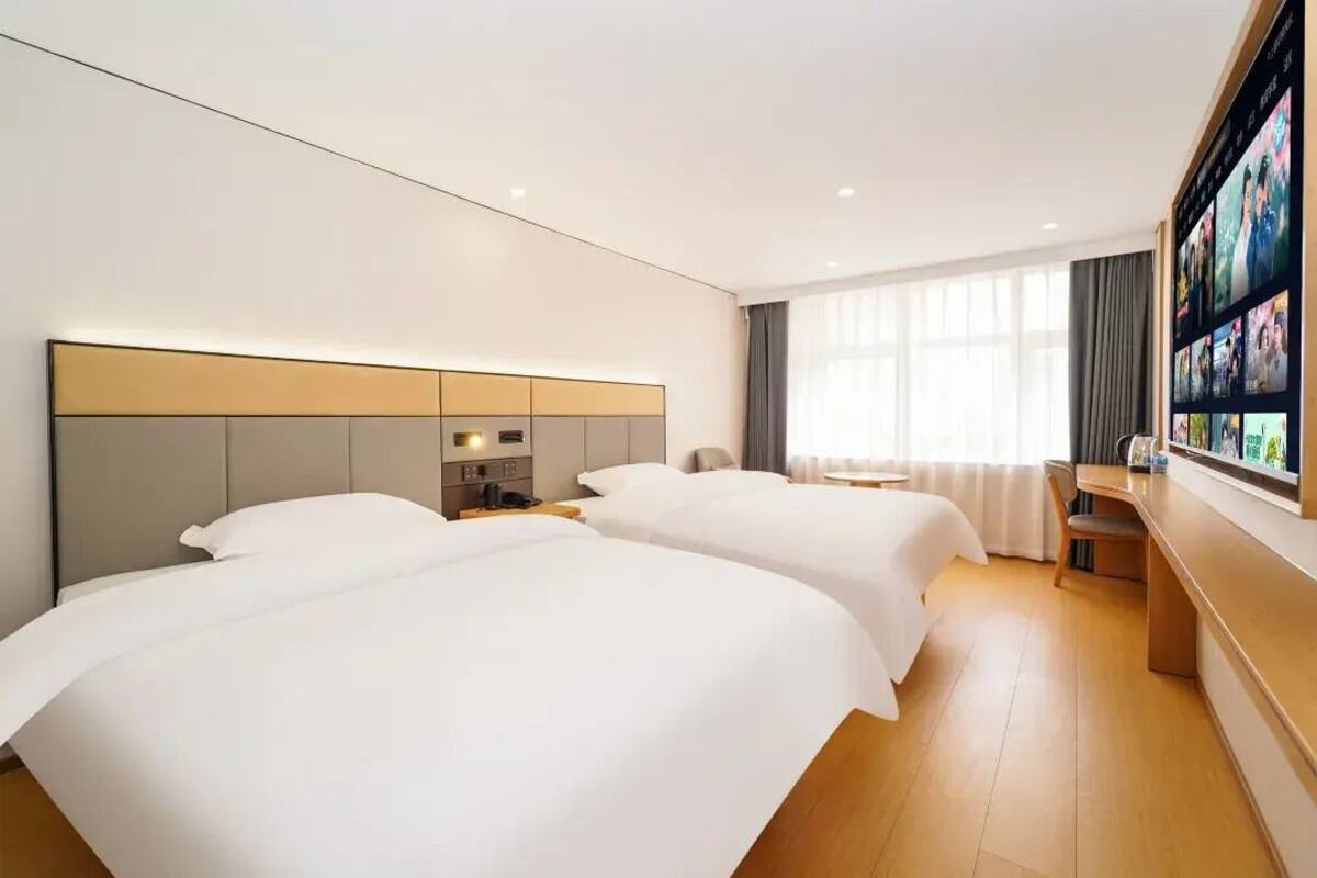 A hotel room with two beds, a desk, a TV mounted on the wall, and a large window with curtains.