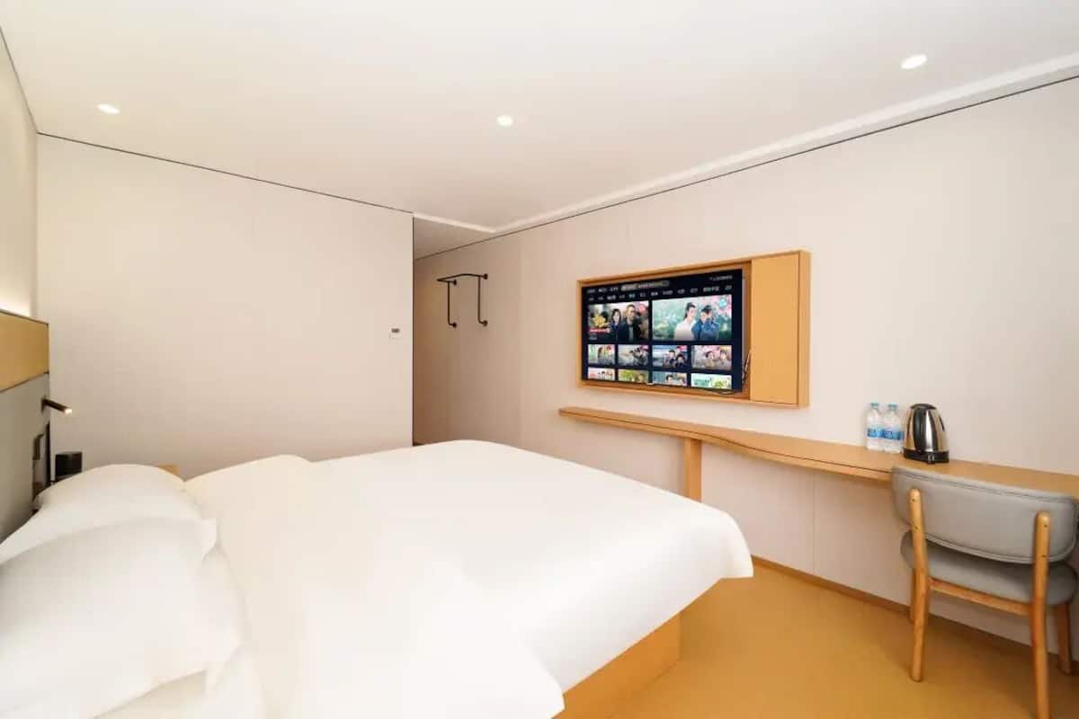 A hotel room with a bed, a desk with a chair, a TV mounted on the wall, and a small shelf with a kettle and bottles.
