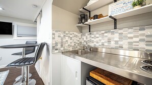 Cozinha compacta com design moderno, destacando uma pia de aço inoxidável, um balcão pequeno e um forno embutido. Há um banquinho de bar e uma mesa com uma cadeira, e uma placa na parede acima do balcão.