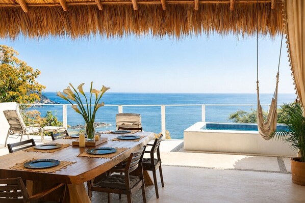 A dining area with a view of the ocean, a pool, and a hanging chair.