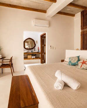 A bedroom with a bed, a wooden bench, a desk, and a bathroom visible through an open door.