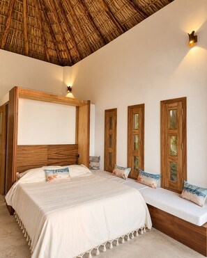 A bedroom with a large bed, wooden headboard, a bench with pillows, and a thatched ceiling.