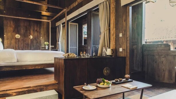 A wooden room with a bed, a dining table with fruits and tea set, and a view of the outdoors. - Tsingpu Tulou Retreat (Zhangzhou)