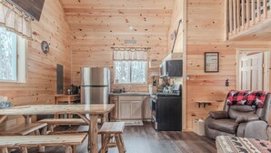 A cabin interior with a wooden dining table, benches, a kitchen area, a living room with a sofa and a recliner, and a window with curtains.