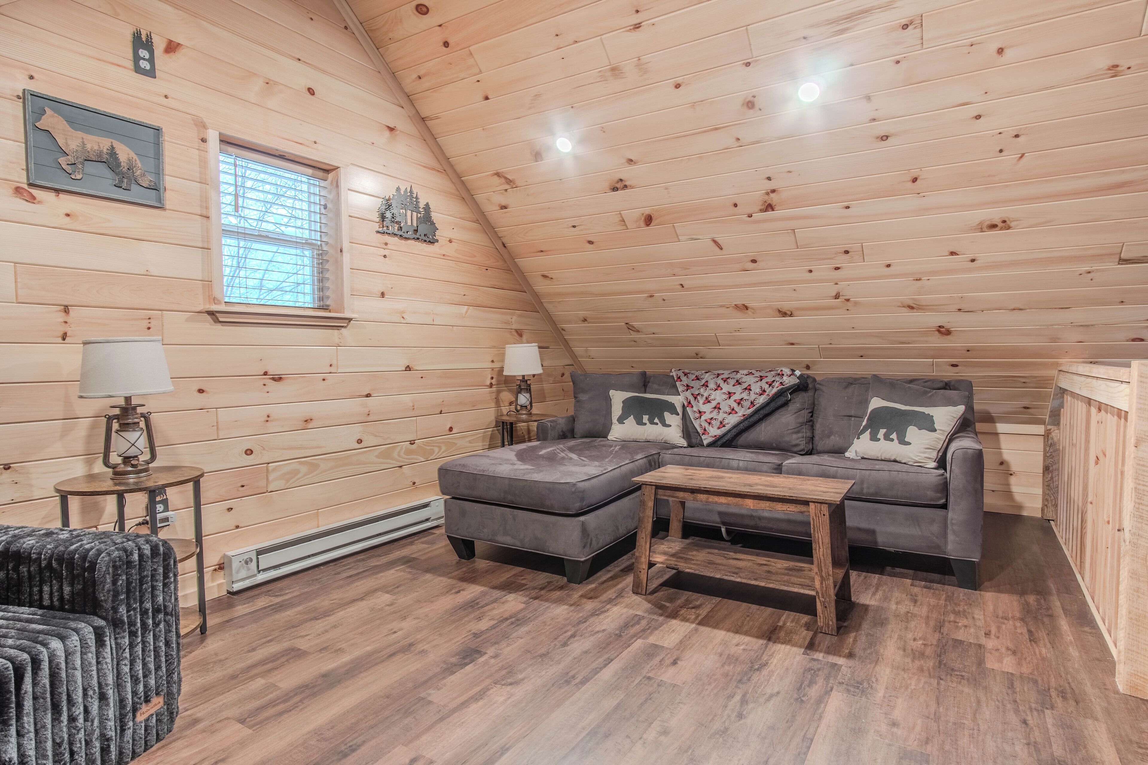 Un acogedor dormitorio tipo loft con techo inclinado, paredes de madera, un sofá gris con almohadones de osos, una mesita y una lámpara.