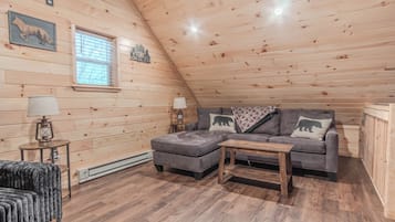 Un acogedor dormitorio tipo loft con techo inclinado, paredes de madera, un sofá gris con almohadones de osos, una mesita y una lámpara.