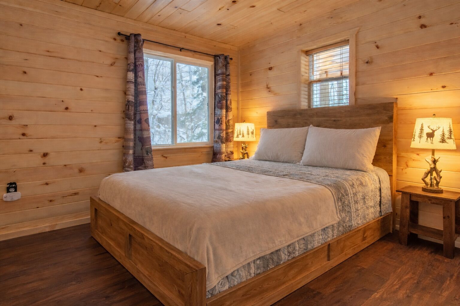 A wooden cabin bedroom with a bed, two windows, and a nightstand with a lamp.