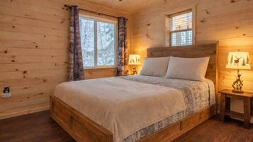 A wooden cabin bedroom with a bed, two windows, and a nightstand with a lamp.
