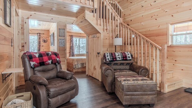 A wooden cabin interior with a brown leather recliner, a matching ottoman, and a staircase.