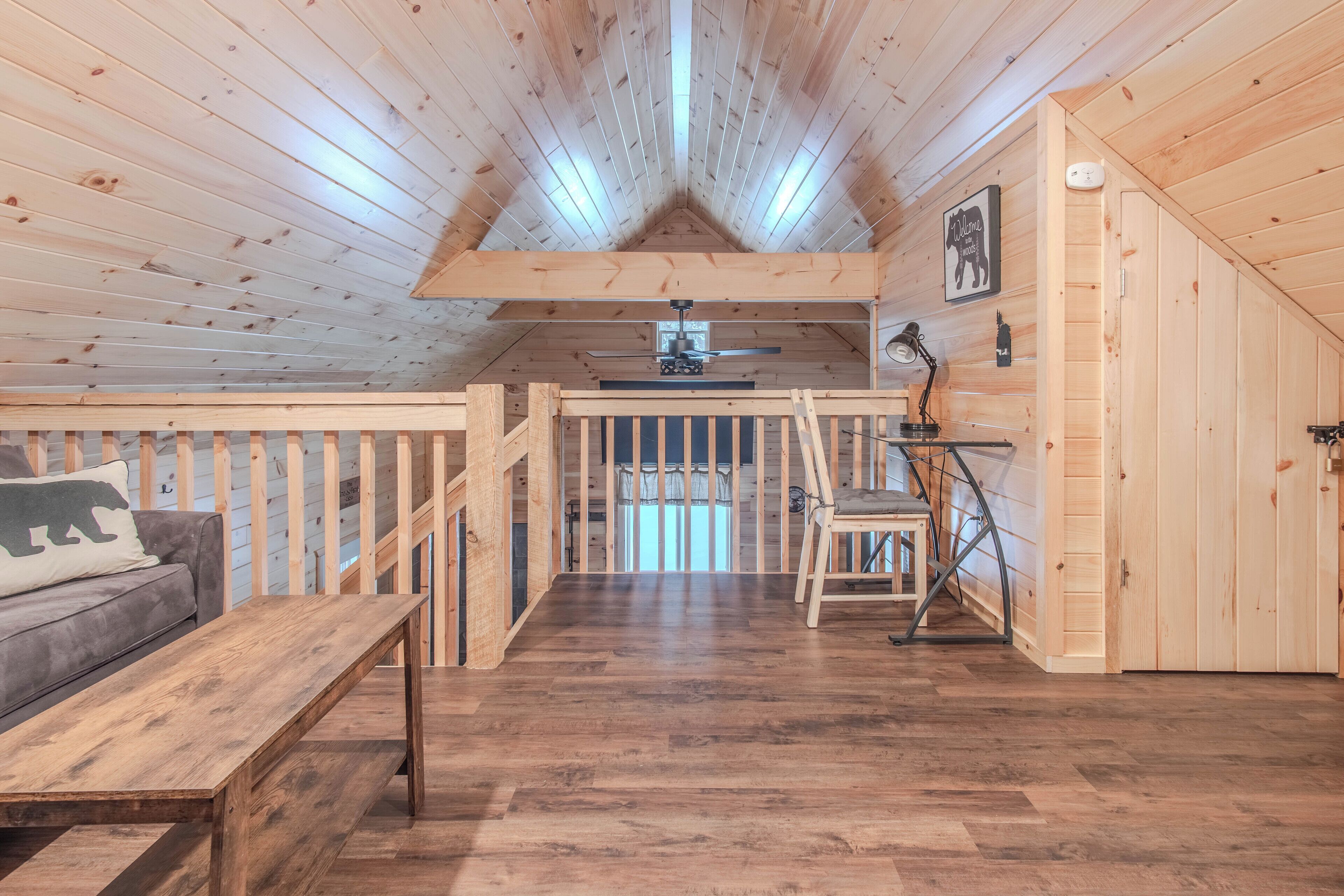 A cozy cabin interior with a wooden ceiling, a lofted sleeping area, a dining table, and a sofa with a bear pillow.