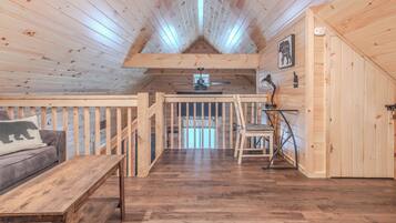A cozy cabin interior with a wooden ceiling, a lofted sleeping area, a dining table, and a sofa with a bear pillow.