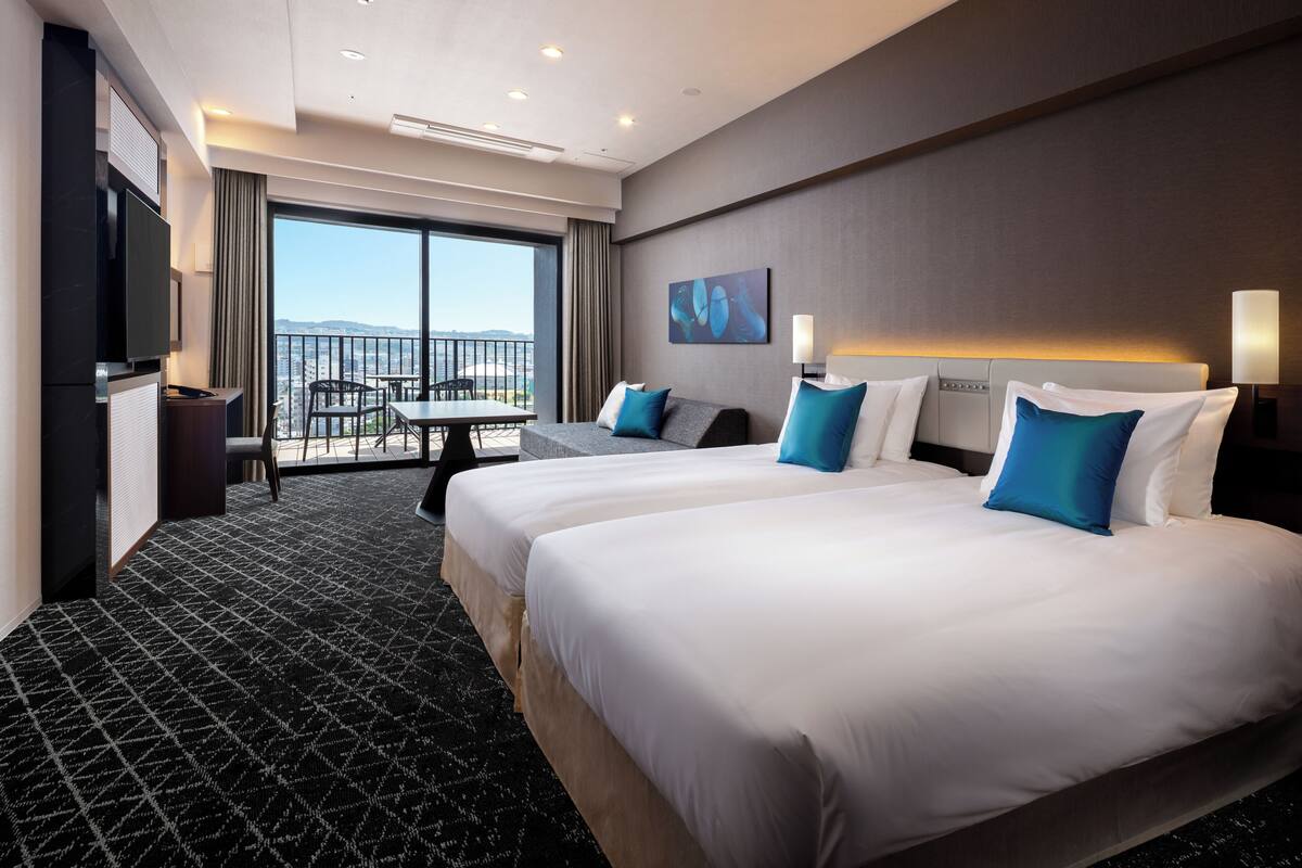 A hotel room with two beds, a TV, a balcony with a table and chairs, and a city view.