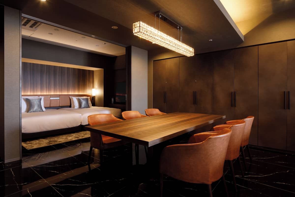A modern hotel room with a large wooden dining table, leather chairs, a sofa bed, and a large flat-screen TV.