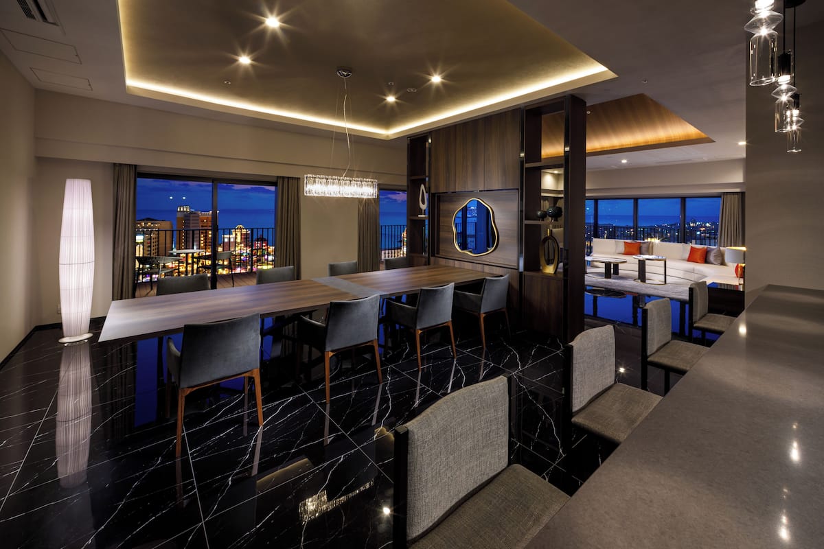 A modern hotel room with a dining area, a bar, and a seating area with a city view.