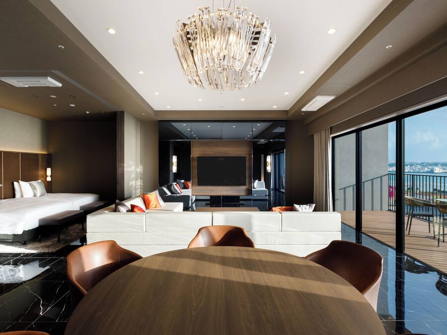 Habitación de hotel moderna con una cama grande, zona de estar, mesa de comedor y balcón con vistas a la ciudad.