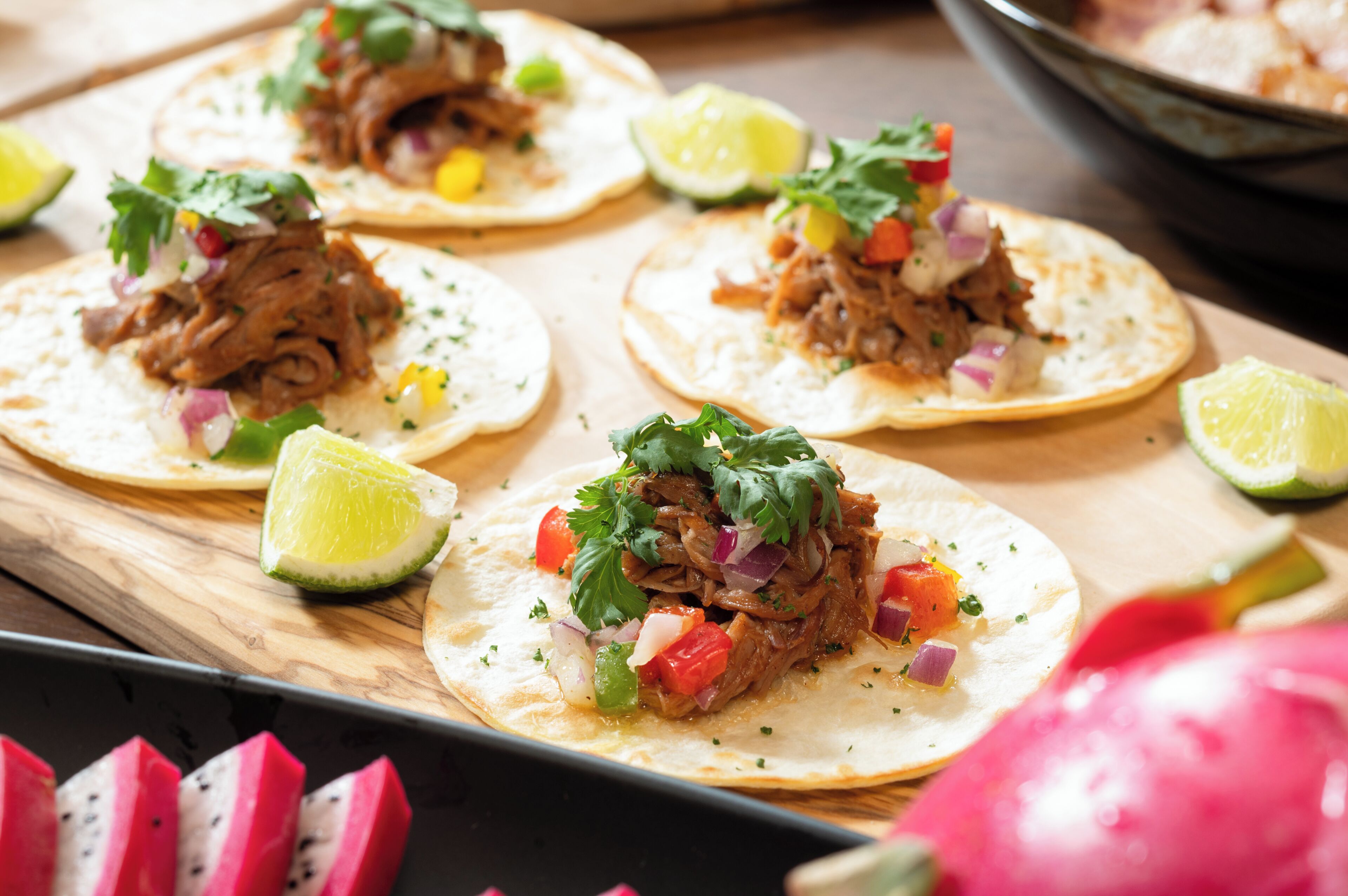 Un plateau de tacos garnis de viande effilochée, de légumes coupés en dés et de quartiers de citron vert.