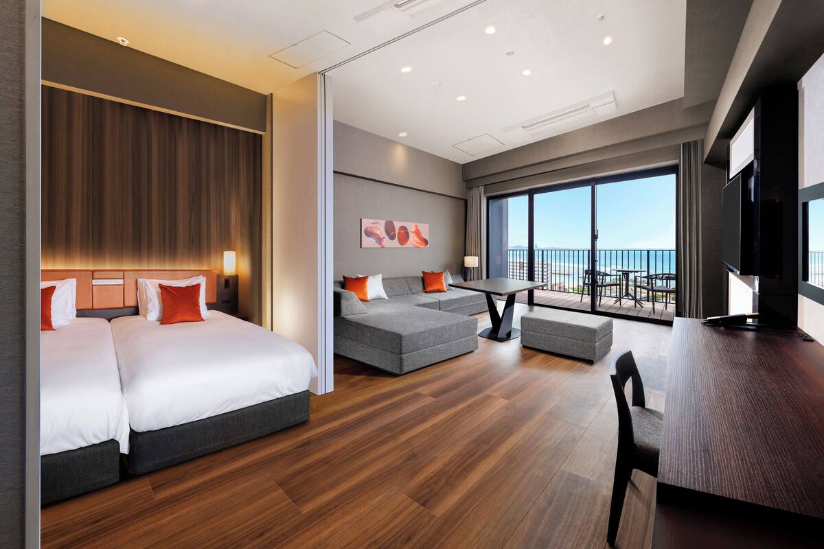 A modern hotel room with a large bed, a sofa, a dining table, and a balcony with a view.