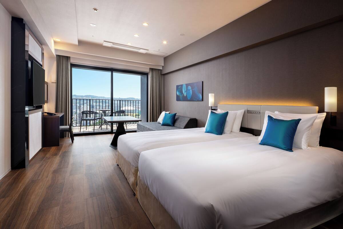 A hotel room with two beds, a TV, a balcony with a table and chairs, and a city view.