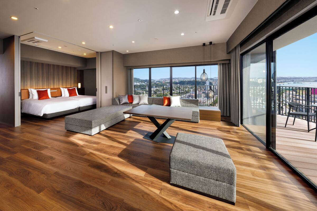A modern hotel room with a large bed, a sofa, a dining table, and a balcony with a view.