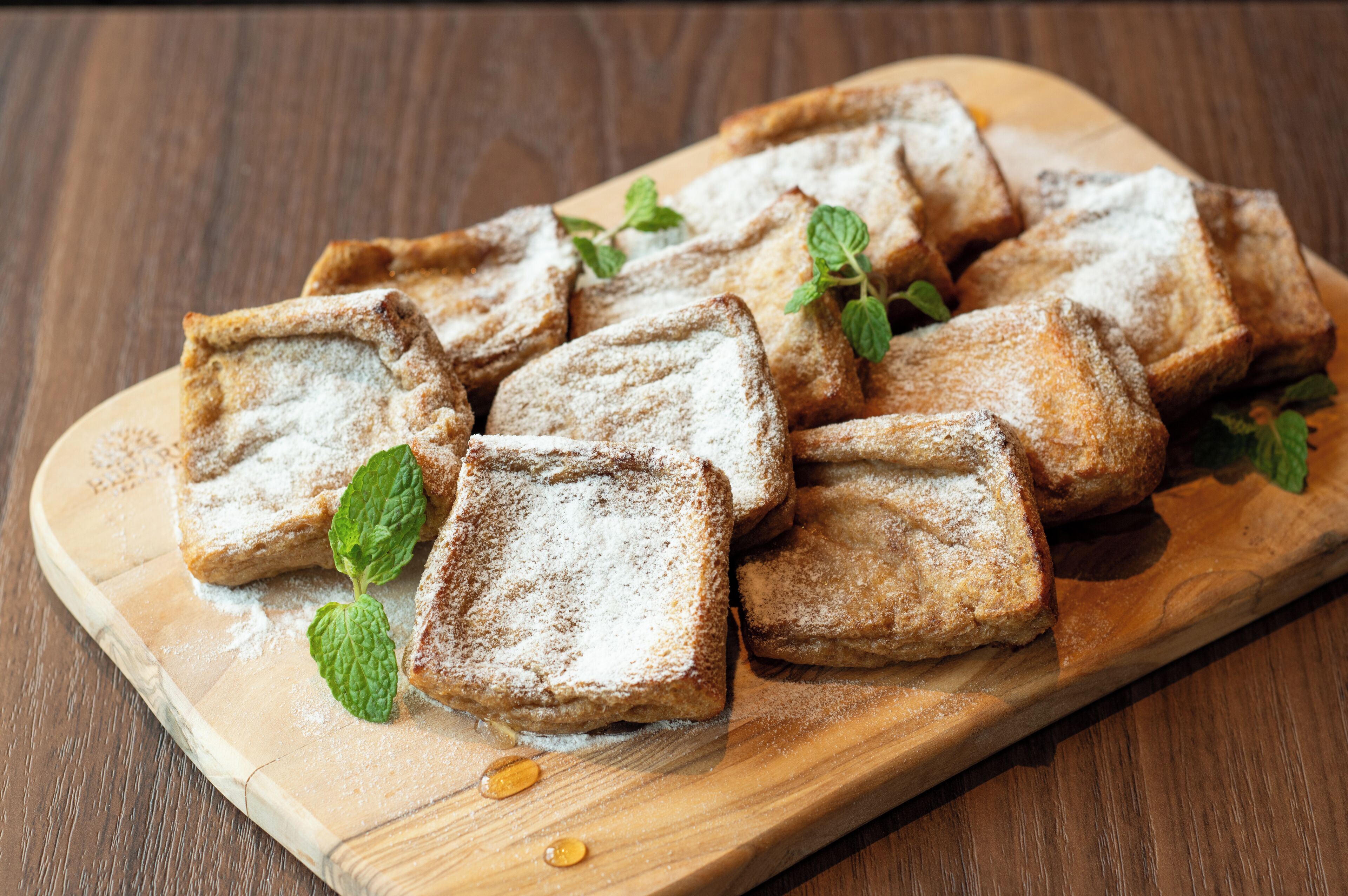A wooden cutting board with several pieces of bread dusted with powdered sugar and garnished with mint leaves.
