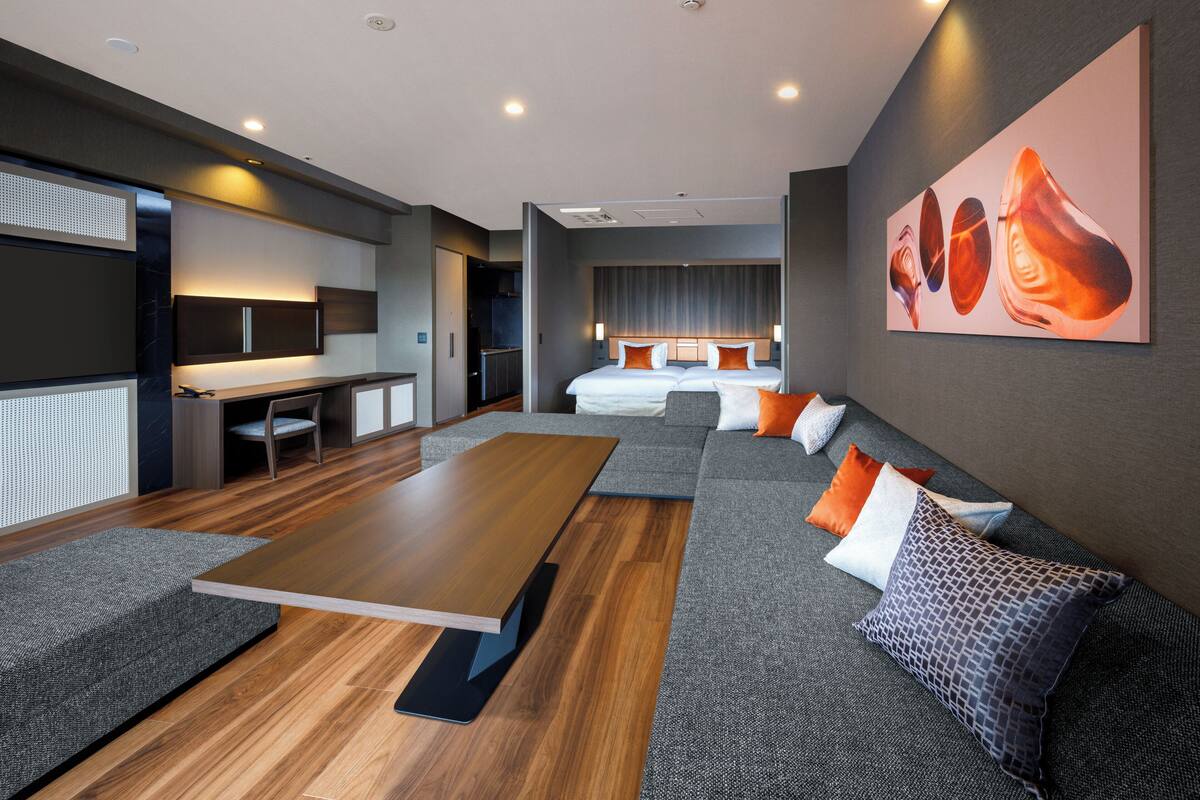 A modern hotel room with a wooden dining table, a sofa with patterned cushions, a bed with white linens, and a large abstract painting on the wall.