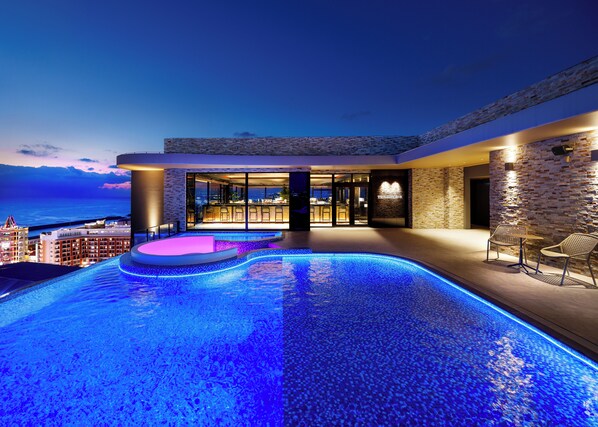 A modern infinity pool with a view of the city at dusk, a bar area with seating, and a stone facade.