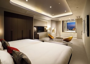 Une chambre d’hôtel moderne, dotée d’un grand lit, d’un bureau, de deux chaises, d’une télévision et offrant une vue sur la ville la nuit.