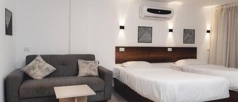 Habitación de hotel con sofá gris, una mesita, cama con cabecera de madera y dos camas individuales.