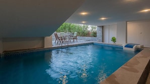 Una piscina interior con borde de azulejos, una pequeña mesa con sillas y vistas a zonas verdes.