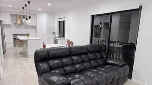Een moderne woonkamer met een zwarte leren bank, een eethoek met een witte tafel en stoelen, en een keuken op de achtergrond.