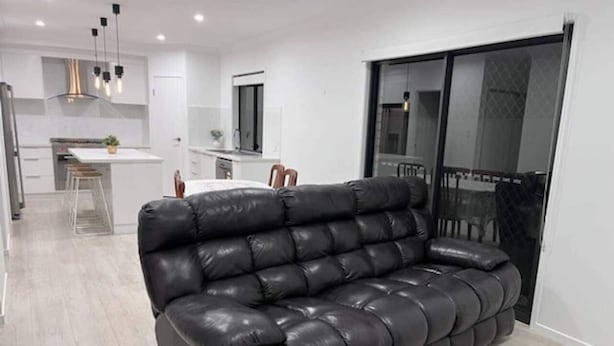Un salon moderne avec un canapé en cuir noir, un coin repas avec une table et des chaises blanches, et une cuisine en arrière-plan.