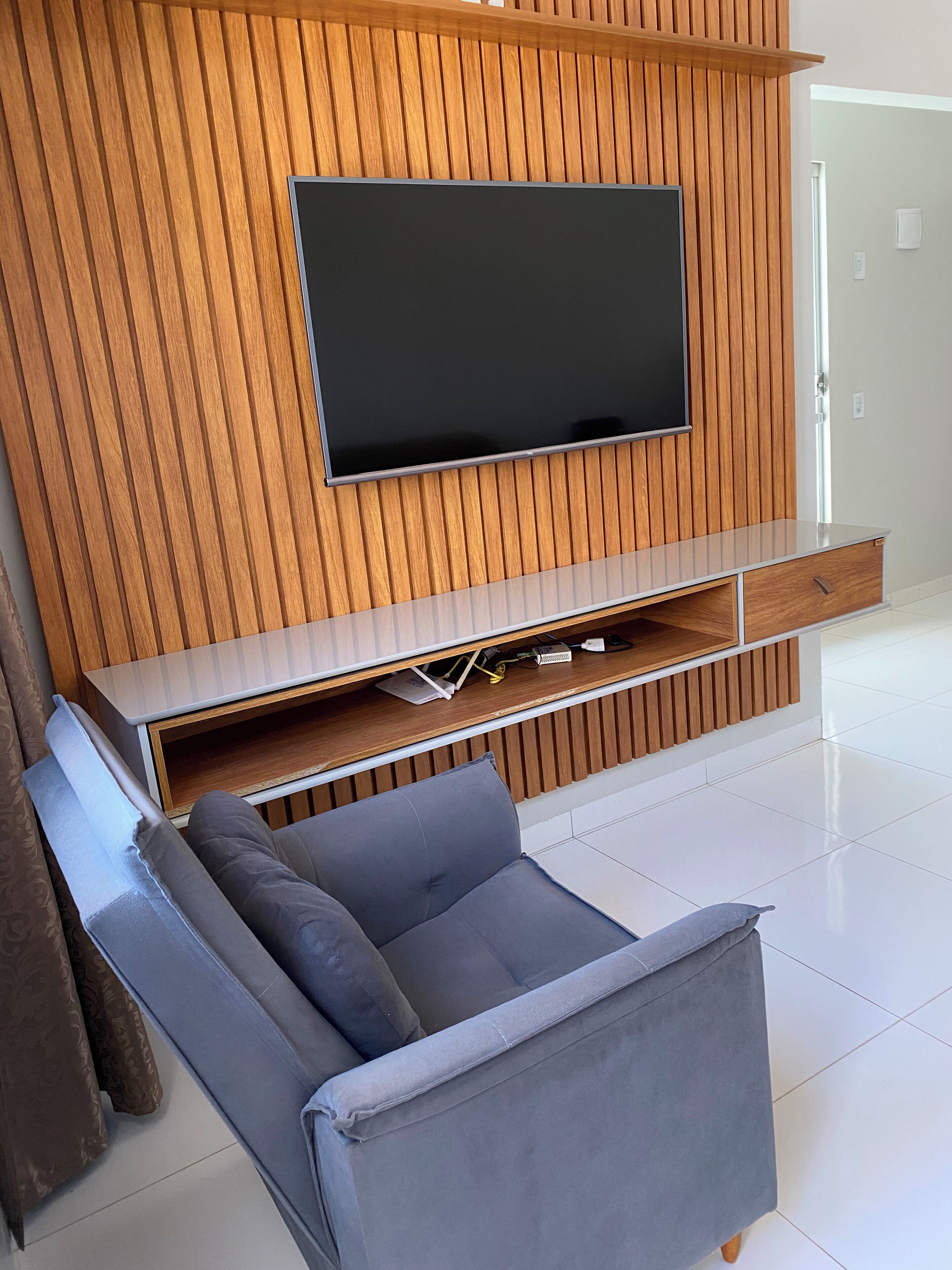 Un salon moderne avec un téléviseur à écran plat fixé à un mur en bois, un canapé gris agrémenté de coussins et un sol carrelé de couleur blanche.