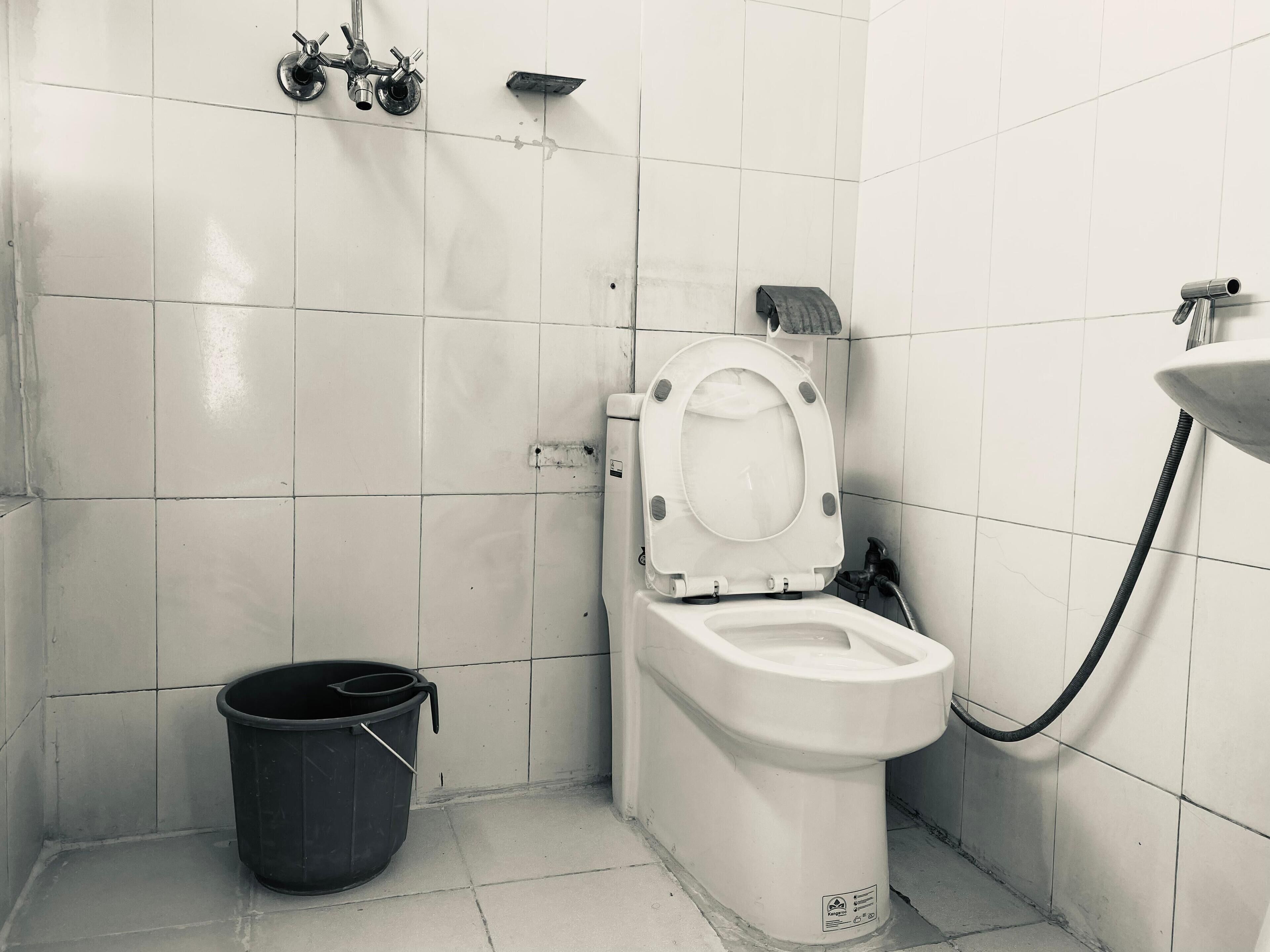Une petite salle de bain carrelée, équipée de toilettes, d’un seau noir et d’un tuyau de douche.