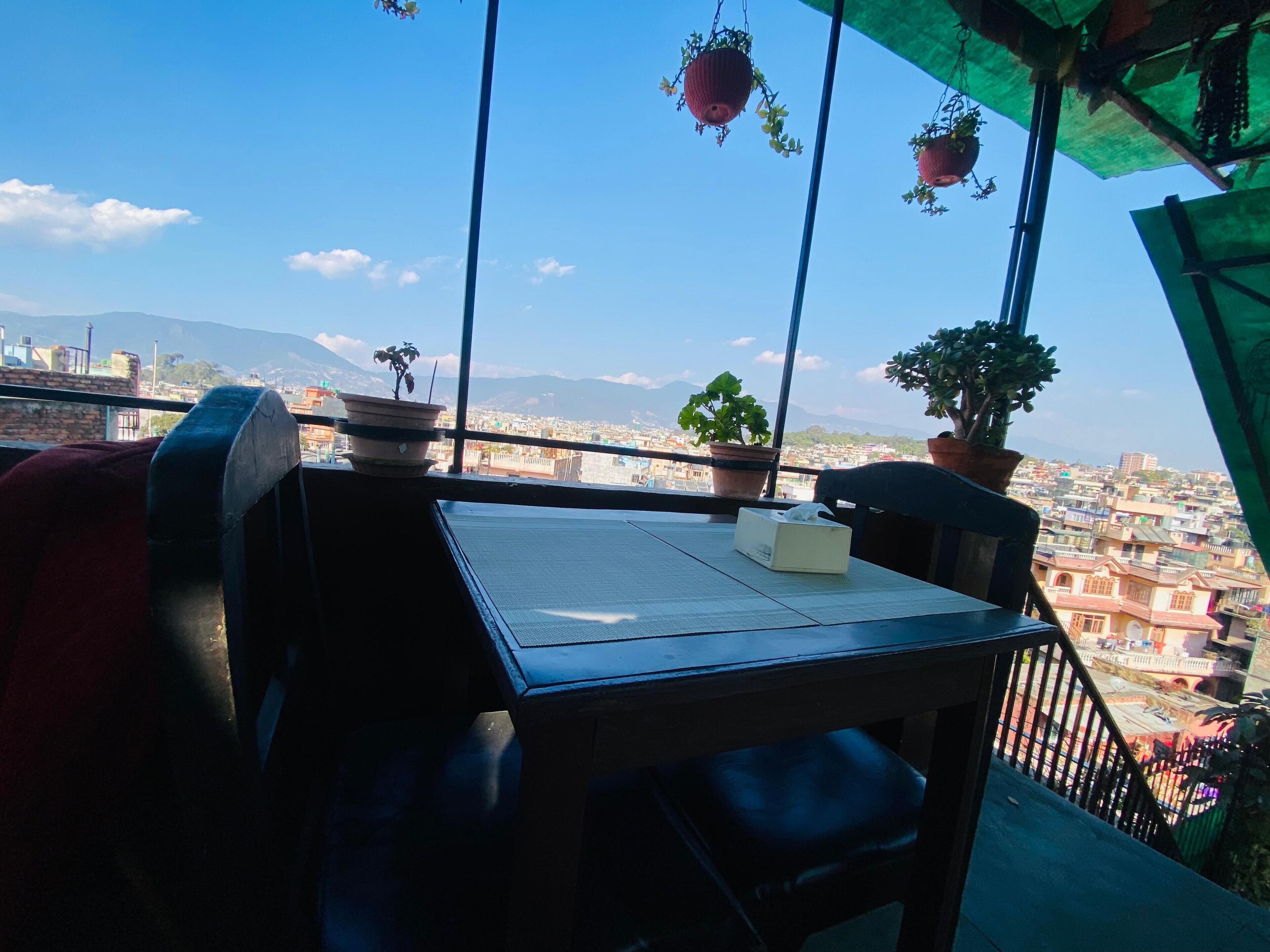 A balcony with a table and chairs, potted plants, and a view of a cityscape with mountains in the distance.