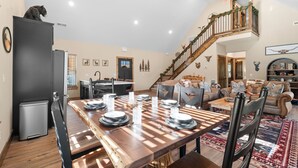 Un salon spacieux avec une table à manger en bois, disposée pour accueillir six personnes, un escalier avec une rampe et une grande fenêtre laissant entrer la lumière naturelle.