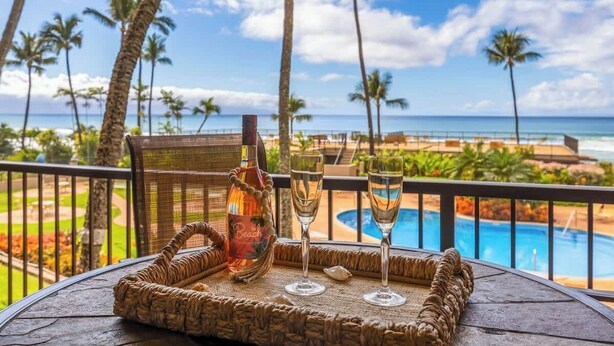 A balcony with a wicker tray, a bottle, and two glasses, overlooking a pool and beach with palm trees and a clear sky.