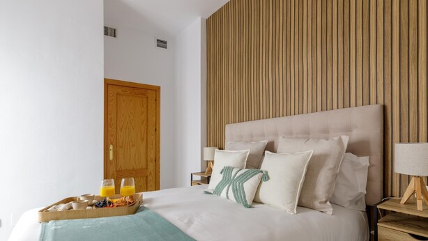 Una cama bien hecha con cabecera de madera, una bandeja con alimentos para el desayuno y una mesita de noche con una lámpara.