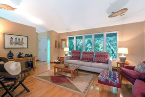 Ein Wohnzimmer mit einer Couch, Sesseln, einem Couchtisch und einem Teppich. Ein Fenster bietet Blick auf Bäume, eine Tür und ein gerahmtes Kunstwerk sind an der Wand zu sehen.