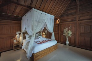 Ein Bett mit Baldachin, ausgestattet mit weißer Bettdecke und herzförmigen Verzierungen, Holzvertäfelung an den Wänden, ein Nachttisch mit Lampe und einer Vase mit Trockenpflanzen sowie eine große Kübelpflanze neben dem Bett.