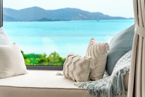 Blick aus einem Fenster mit einer Couch und Kissen auf ein Gewässer und entfernte Berge.