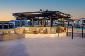 Un área de estar al aire libre con techo, sillones reclinables y una mesa, rodeada de macetas de concreto y una piscina.