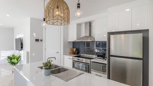 A modern kitchen with white cabinetry, stainless steel appliances, a large island with a sink, and a unique woven pendant light fixture. - Lavender & Olive - Pet Friendly, Family Friendly (Myrtleford)